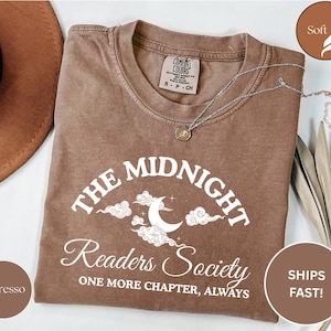 Könnte beinhalten: Espressofarbenes T-Shirt mit dem weißen Text "THE MIDNIGHT Readers Society". Das Design umfasst einen Mond, Wolken und Sterne. Eine silberne Halskette mit Anhänger liegt auf dem Shirt. Das Shirt ist mit "Comfort Colors" gekennzeichnet.