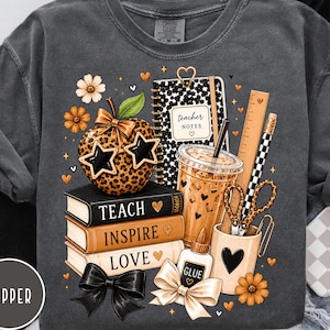 May include: A charcoal grey t-shirt featuring a graphic print of a leopard print apple wearing star-shaped sunglasses, a notebook labelled teacher notes, a stack of books reading teach, inspire, love, a coffee cup, a ruler, scissors, and glue.