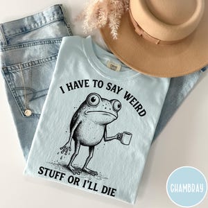 May include: Light blue t-shirt with a black and white illustration of a frog holding a mug. The text on the shirt reads "I HAVE TO SAY WEIRD STUFF OR I'LL DIE". The shirt is paired with blue jeans and a tan hat.