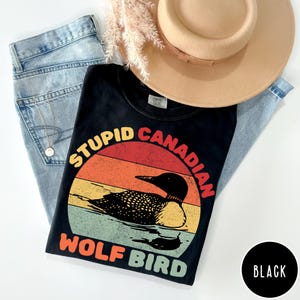 Pode incluir: Camiseta preta com um gráfico de um mergulhão num design de pôr do sol. O texto na camisa diz "STUPID CANADIAN WOLF BIRD". A camisa é combinada com jeans azul claro e um chapéu bege.