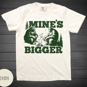 Puede incluir: Una camiseta blanca con un gráfico verde de dos osos sentados junto a una fogata. El texto "Mine's Bigger" está impreso en verde debajo de los osos.