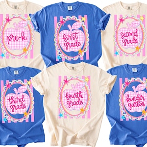 Könnte beinhalten: Sechs kurzärmelige T-Shirts in Creme und Blau, jedes mit einer zentralen Grafik eines rosafarbenen Vichy-Apfels in einem dekorativen ovalen Rahmen. Der Text in den Äpfeln lautet pre-k, first grade, second grade, third grade, fourth grade und kindergarten.