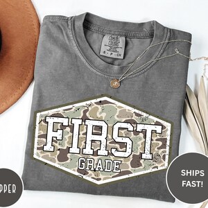 以下が含まれることがあります： チャコールグレーの半袖Tシャツ。迷彩柄の六角形のグラフィックと、白い文字で「FIRST GRADE」とプリントされています。クルーネックで、襟の内側にはComfort Colorsのブランドラベルがプリントされています。