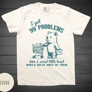 May include: A white t-shirt featuring a teal graphic of a bear pushing a shopping trolley full of items. The text "I got 99 problems and a sweet little treat would solve most of them" is printed below the bear.