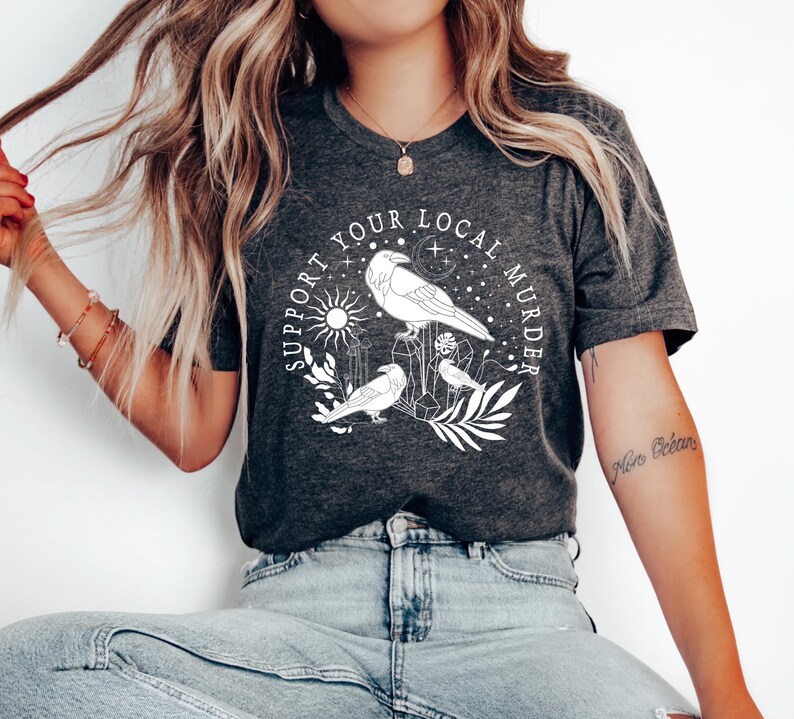 May include: A dark gray t-shirt with a white graphic design that says "Support Your Local Murder" with a raven and other birds, stars, and crystals.