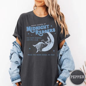 Op de afbeelding: Donkergrijs T-shirt met de tekst "secret society of MIDNIGHT READERS" in blauw. Het shirt heeft een afbeelding van een vrouw die op een maan leest, met de tekst "One more chapter, now it's 3 A.M." en "While the world sleeps, we read".