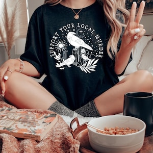 May include: A black t-shirt with a white graphic design featuring a circle with the text "Support Your Local Murder" and three stylized ravens with a crescent moon, stars, and sun.