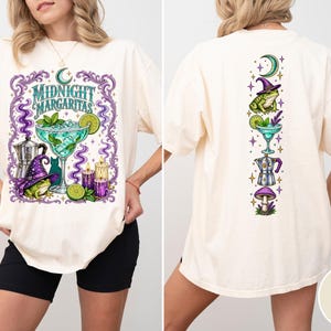 Könnte beinhalten: Ein elfenbeinfarbenes T-Shirt mit einem Grafikdruck und dem Text Midnight Margaritas. Das Vorderdesign zeigt ein Margaritaglas, einen Frosch mit violettem Hexenhut, eine Kaffeekanne, Kerzen und eine Katze. Die Rückseite zeigt vertikal angeordnete Symbole.