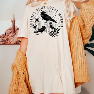 May include: A white t-shirt with a black graphic design featuring crows, a crescent moon, a sun, and the text "Support Your Local Murder".