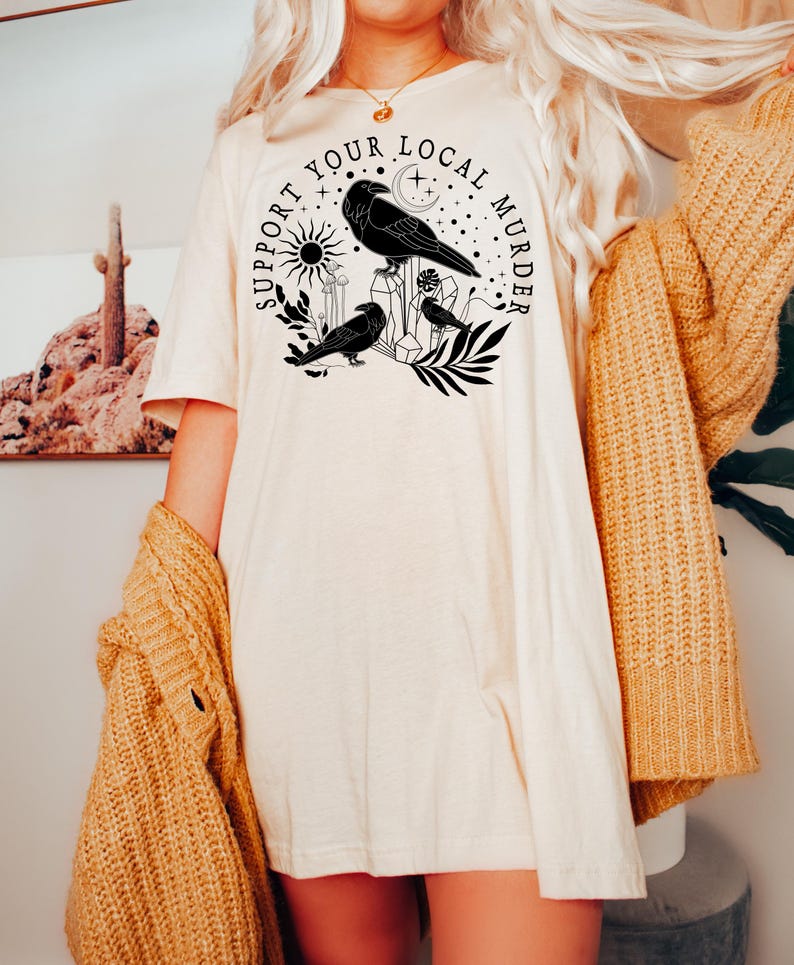 May include: A white t-shirt with a black graphic design featuring crows, a crescent moon, a sun, and the text "Support Your Local Murder".