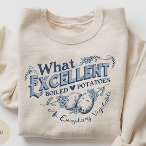 Puede incluir: Una sudadera beige con un diseño gráfico que dice "What Excellent Boiled Potatoes An Exemplary Vegetable". El gráfico presenta un corazón, dos patatas y flores azules.