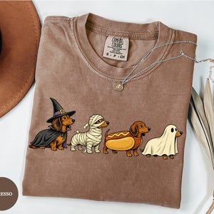 May include: An espresso-colored t-shirt featuring a graphic of four dachshunds in Halloween costumes: one as a witch, one as a mummy, one as a hot dog, and one as a ghost. The shirt has a Comfort Colors label at the neck and a crew neckline.