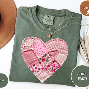 May include: A moss green t-shirt with a patchwork heart design in shades of pink and floral patterns. A delicate gold necklace rests on the shirt. The shirt has a tag that says "Comfort Colors".