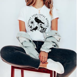 May include: A white t-shirt with a black graphic design featuring three crows, a crescent moon, a sun, and the text "Support Your Local Murder".