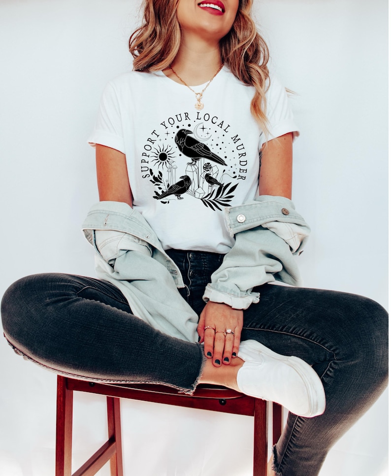 May include: A white t-shirt with a black graphic design featuring three crows, a crescent moon, a sun, and the text "Support Your Local Murder".