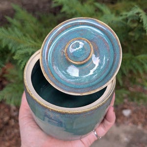May include: A round ceramic jar with a matching lid, featuring a glossy teal and blue glaze with brown rim accents. The jar has a smooth, cylindrical body and a small circular handle on the center of the lid, showcasing a handcrafted finish.
