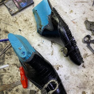 May include: Two black leather shoes in the process of being made, with blue shoe forms inside. The shoes have a crocodile skin texture, silver buckles, and are held together with small pins. The shoes are on a work surface with tools.