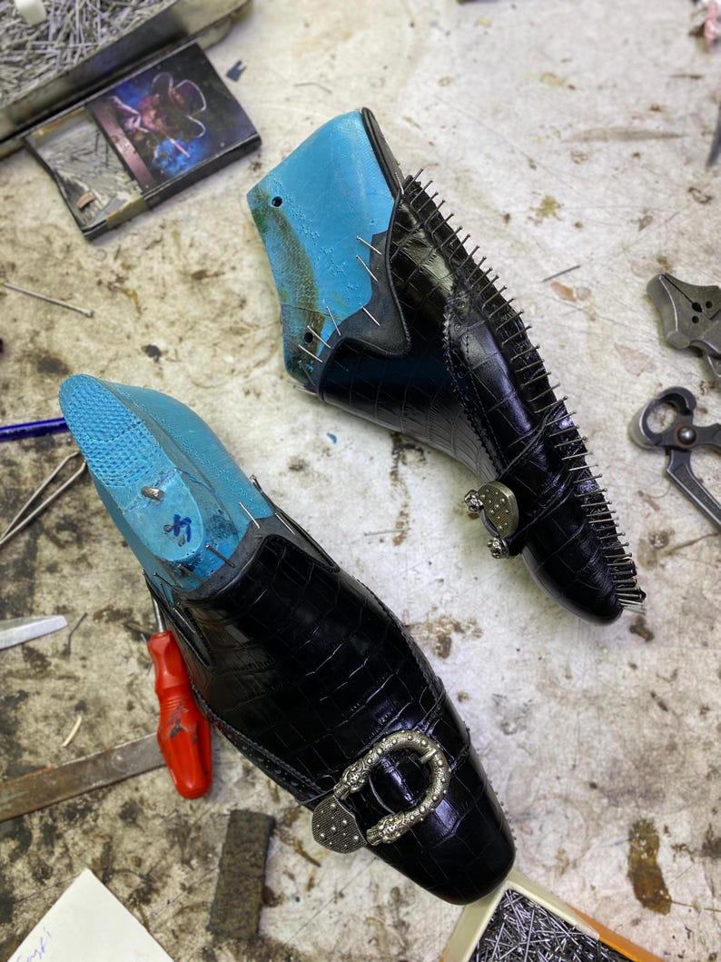 May include: Two black leather shoes in the process of being made, with blue shoe forms inside. The shoes have a crocodile skin texture, silver buckles, and are held together with small pins. The shoes are on a work surface with tools.