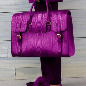 May include: A vibrant purple handbag with a snakeskin texture, featuring gold-colored buckles and a top handle. The bag is held by a person wearing matching purple pants and slippers with fur trim. The overall aesthetic is bold and luxurious.