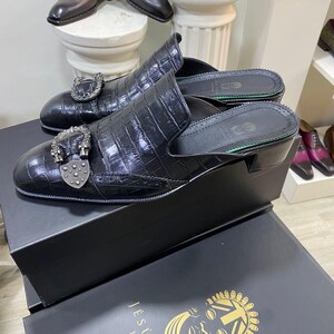 May include: Black leather mules with a crocodile texture and decorative silver buckles. The shoes are displayed on a black box with the text "JESU-S&Eacute;G LONDON". Additional leather shoes are visible in the background.