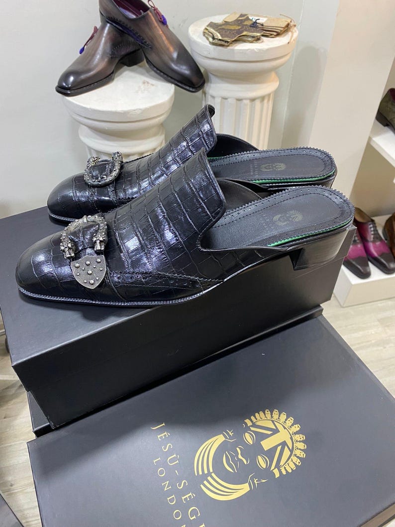May include: Black leather mules with a crocodile texture and decorative silver buckles. The shoes are displayed on a black box with the text "JESU-S&Eacute;G LONDON". Additional leather shoes are visible in the background.