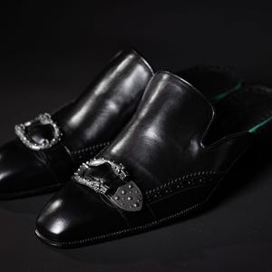 May include: A pair of black leather slip-on shoes with silver-colored decorative buckles. The shoes have a square toe and a green insole. The background is black.
