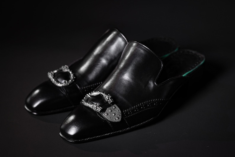 May include: A pair of black leather slip-on shoes with silver-colored decorative buckles. The shoes have a square toe and a green insole. The background is black.