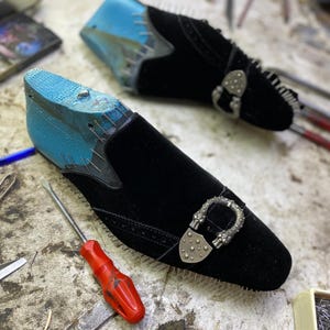 May include: Black velvet slip-on shoes with silver buckles, in the process of being made. The shoe forms are blue. A red screwdriver and other tools are on the work surface.