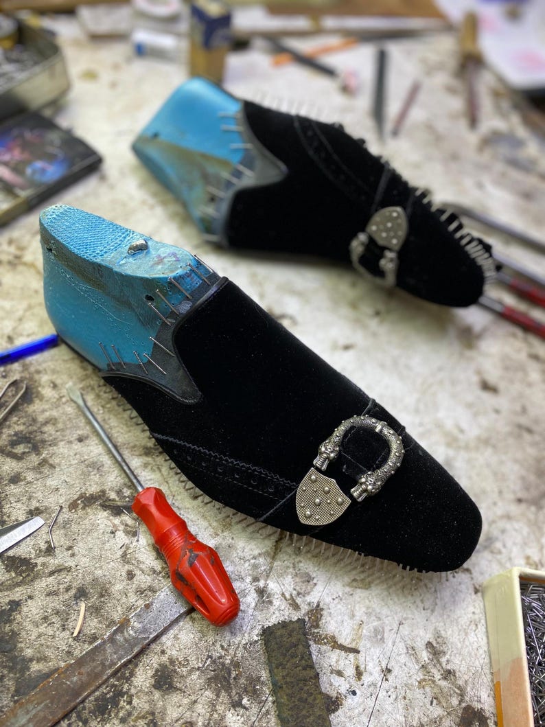 May include: Black velvet slip-on shoes with silver buckles, in the process of being made. The shoe forms are blue. A red screwdriver and other tools are on the work surface.