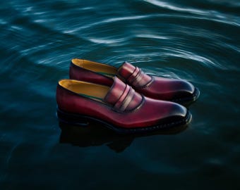 May include: A pair of burgundy leather loafers with a classic penny loafer design. The shoes have a polished finish and are set against a dark blue water background, creating a striking contrast.