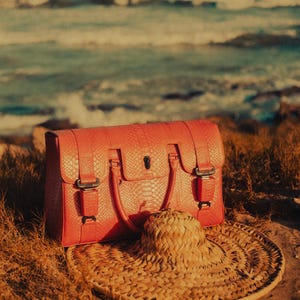 Puede incluir: Un bolso de mano de color coral con un patrón de piel de serpiente texturizado, con un asa superior y una solapa frontal con hebillas. Un sombrero de paja para el sol descansa frente al bolso. El fondo muestra el mar y la hierba de la playa.