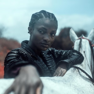 Könnte beinhalten: Eine Person mit dunkler Haut und geflochtenem Haar, die eine schwarze, strukturierte Jacke trägt, lehnt sich an ein weißes Pferd. Der Hintergrund zeigt einen blauen Himmel und verschwommenes Laub, was ein dramatisches Porträt ergibt.