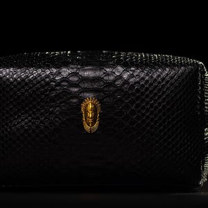 May include: Black rectangular cosmetic bag with a textured snakeskin pattern. The bag features a gold-colored emblem and a small handle. The bag is set against a black background.