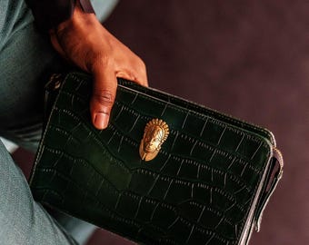 May include: Dark green clutch with a crocodile texture. The clutch features a gold-colored decorative emblem of a face. The bag has a zipper closure and is held by a person wearing a black shirt and blue jeans.
