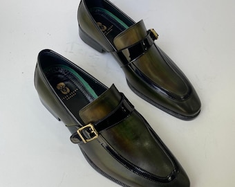 May include: A pair of green leather loafers with a glossy black strap and gold buckle. The shoes have a classic design with a rounded toe and a low heel. The leather has a subtle, aged appearance.