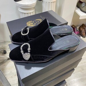 May include: Black velvet slip-on shoes with silver-tone hardware. The shoes have a low heel and are displayed on a black box. Other shoes are visible in the background.