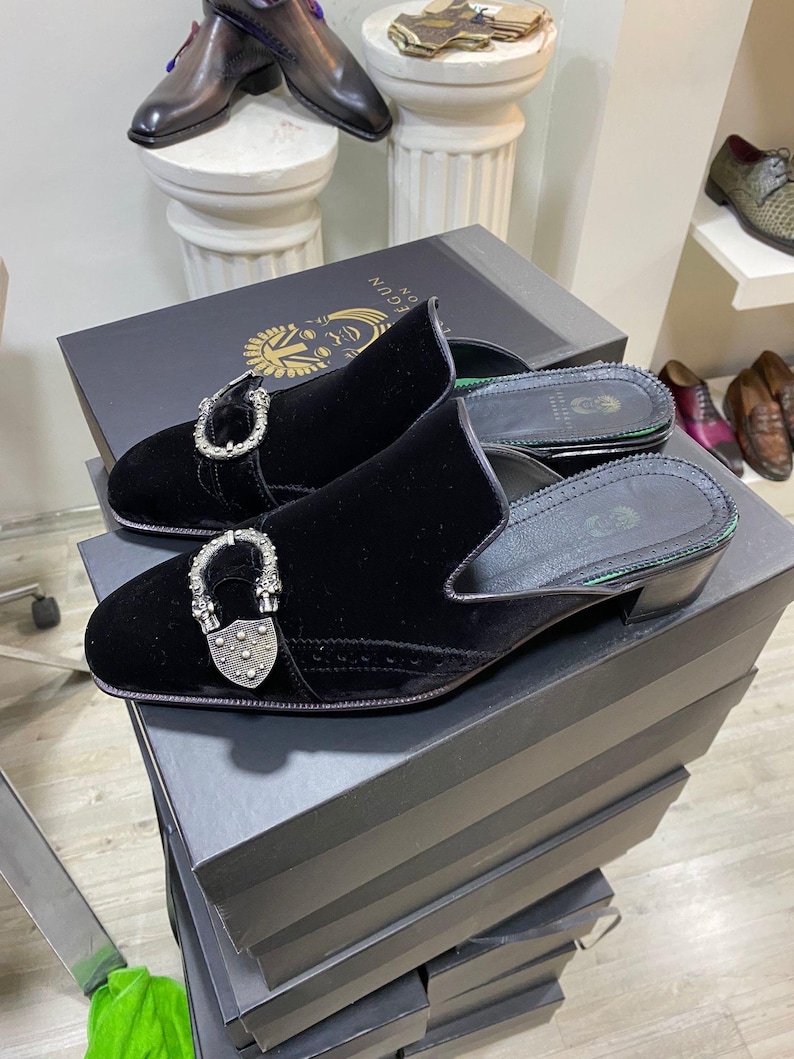 May include: Black velvet slip-on shoes with silver-tone hardware. The shoes have a low heel and are displayed on a black box. Other shoes are visible in the background.