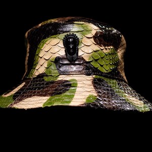 May include: A camouflage-patterned bucket hat with a black decorative element. The hat features a mix of green, brown, and beige tones, with a textured, scaly appearance. The black decorative element is a sculpted face.