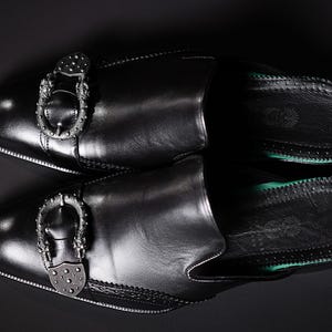 May include: A pair of black leather slip-on shoes with decorative silver buckles. The shoes have a rounded toe and a green sole. The image is shot from above against a dark background.