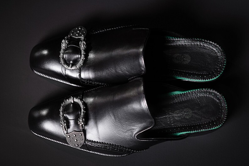 May include: A pair of black leather slip-on shoes with decorative silver buckles. The shoes have a rounded toe and a green sole. The image is shot from above against a dark background.