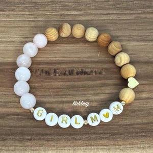 May include: A beaded bracelet featuring a mix of light pink and wooden beads. The bracelet includes white beads with the gold letters spelling "GIRL MOM" and a small gold heart charm. The word "Kahleaj" is also visible.