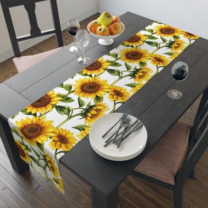 May include: A white table runner with a sunflower pattern. The runner is on a dark wood table with two place settings, each with a white plate and silverware. There are two glasses of wine on the table, and a bowl of apples in the background.
