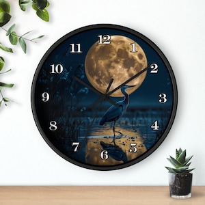 May include: Round black wall clock featuring a serene scene of a heron standing in water under a full moon. The clock face has black hands and white numerals. The clock is a decorative item for home decor.