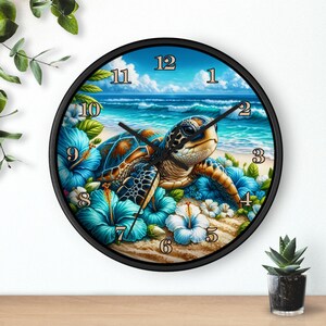 May include: A round wall clock with a black frame featuring a sea turtle on a sandy beach with blue hibiscus flowers. The background shows a blue ocean and sky. The clock hands are black.