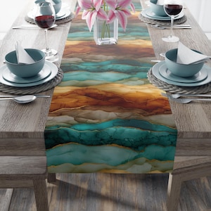 May include: A table runner with an abstract design in shades of teal, brown, and gold. The runner is placed on a wooden table set for a meal, with plates, bowls, silverware, and wine glasses. A vase of pink flowers sits in the center.