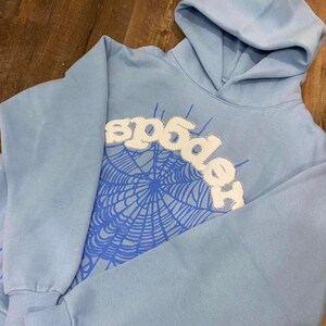 May include: A light blue hoodie with a hood and long sleeves. The front features a white stitched design that spells "spider" above a blue spiderweb graphic. The hoodie is made of a soft material.