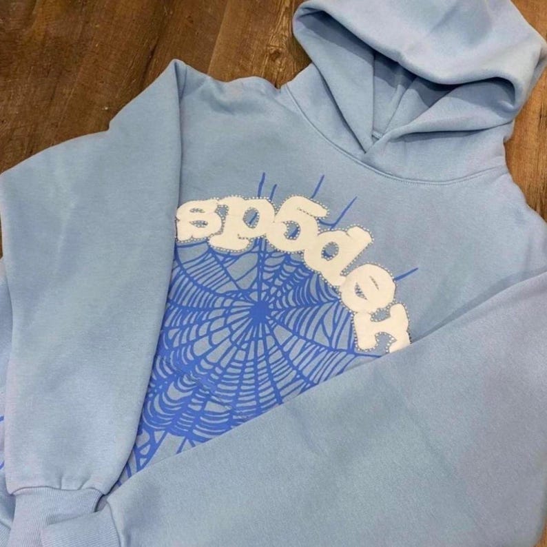 May include: A light blue hoodie with a hood and long sleeves. The front features a white stitched design that spells "spider" above a blue spiderweb graphic. The hoodie is made of a soft material.