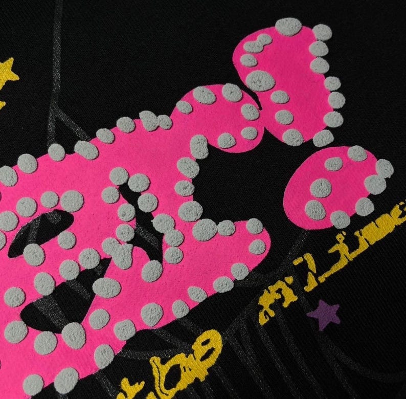 May include: Black fabric with a large, textured pink graphic and gray dots. Yellow and purple accents are visible, along with the partial text "TIME". The design has a distressed, vintage aesthetic.