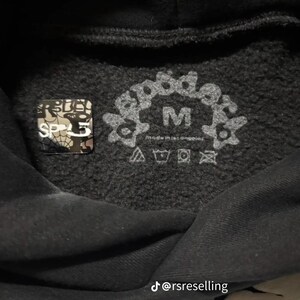 Puede incluir: Primer plano del interior de una sudadera con capucha negra con una etiqueta de talla M y s&iacute;mbolos de cuidado. La etiqueta presenta el texto "good design" en un dise&ntilde;o floral. Tambi&eacute;n es visible una peque&ntilde;a pegatina cuadrada con el texto "SP5".