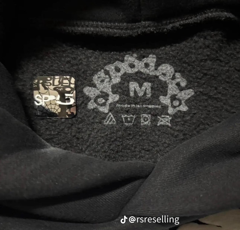 Puede incluir: Primer plano del interior de una sudadera con capucha negra con una etiqueta de talla M y s&iacute;mbolos de cuidado. La etiqueta presenta el texto "good design" en un dise&ntilde;o floral. Tambi&eacute;n es visible una peque&ntilde;a pegatina cuadrada con el texto "SP5".